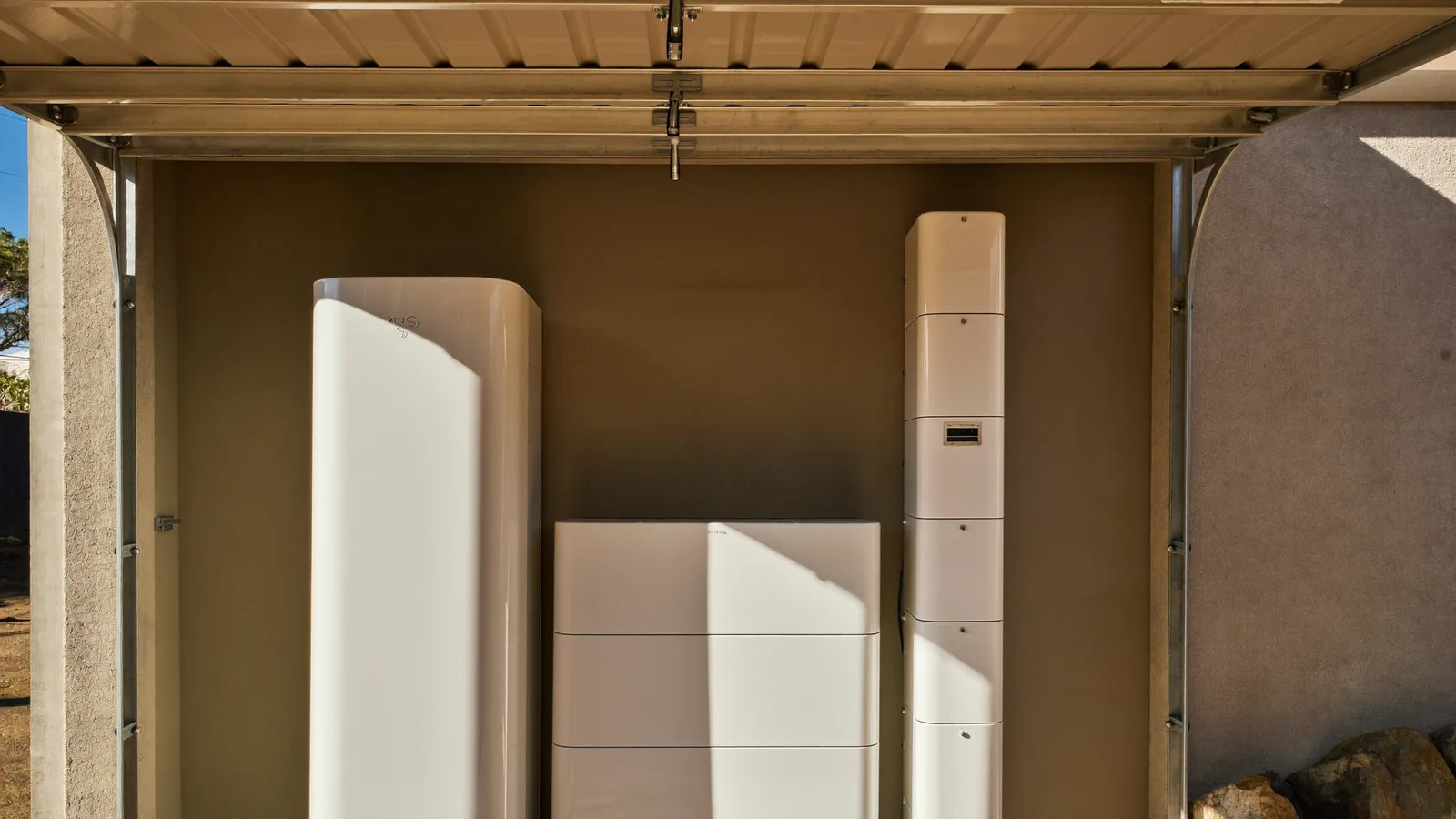 Three home batteries of different designs lined up along a garage wall