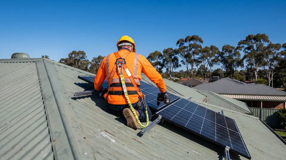 An installer in a safety harness fitting solar panels on a tin roof